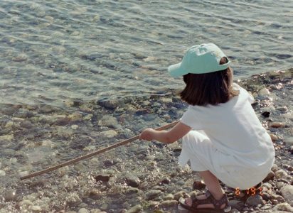 A young girl squatting down on a rocky shoreline using a stick to poke at the water's edge.