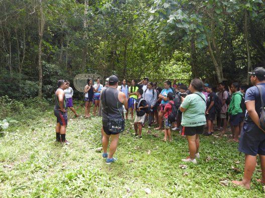 A local school group participates in a guided tour at the Takitumu Conservation Area, an OECM in Takitumu District, Cook Islands, managed by the local community. A group of students and adults stand in a lush, green forest clearing as two guides speak to them. A sign reading 'Welcome to the Takitumu Conservation Area' is visible in the background.