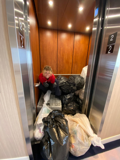 A deckhand in the process of hauling the ships trash for the week off of the ship. The deckhand is perched on top of a pile of trash bags that are toppling out of the elevator.