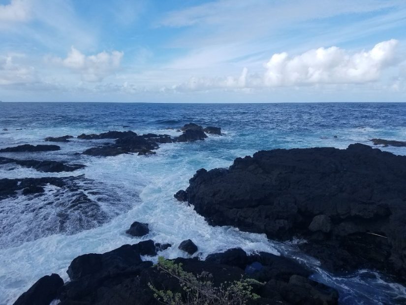 A rocky intertidal coastline with blue foamy water crashing into dark gray volcanic rock.