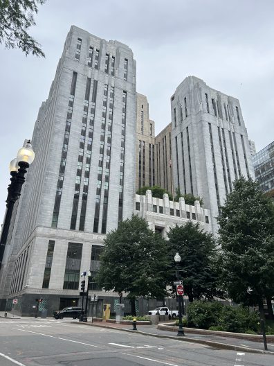 A tall government building with a partial green roof reaches towards a gray sky. A small park and city street lie in the foreground.