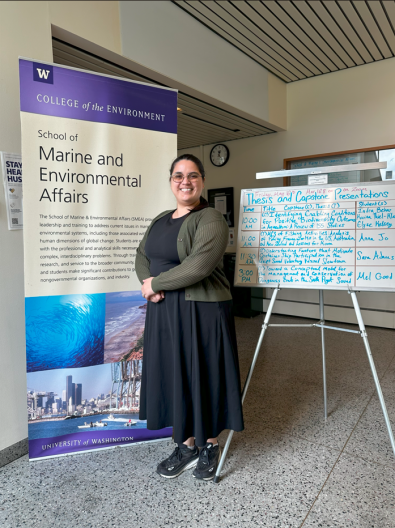 A person in a black dress and green sweater stands smiling next to a "School of Marine and Environmental Affairs" sign and a presentation schedule on a whiteboard.