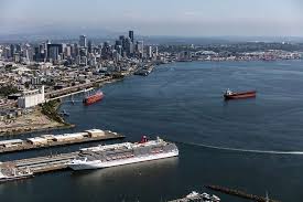 Aerial view of the Seattle waterfront showing downtown skyscrapers, several cargo ships in Elliott Bay, and a large cruise ship docked at a pier in the foreground.