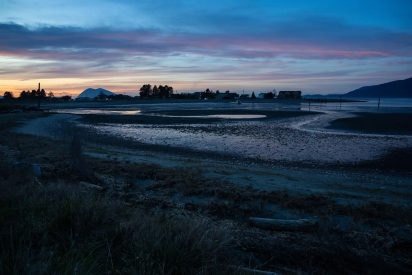 Sunset over a tranquil coastal landscape with calm waters reflecting the evening sky. Silhouetted trees and distant hills create a serene mood.