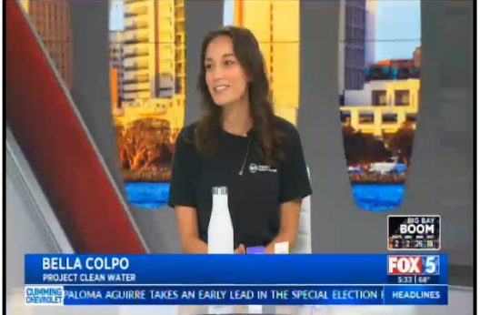 A woman wearing a black "Project Clean Water" shirt smiles during a TV interview. A cityscape backdrop and "FOX 5" branding are visible.
