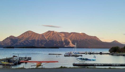 A tranquil lake scene at sunset with mountains in the background. A docked seaplane and boats sit in the foreground, creating a peaceful, serene atmosphere.