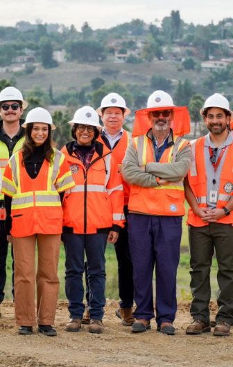 A group of six people wearing orange safety vests and white hard hats stand on a dirt path outdoors, smiling confidently. The background shows hills and trees, suggesting a construction or survey site.
