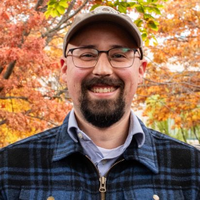 A smiling man in glasses and a cap stands outdoors with vibrant autumn leaves in the background, wearing a blue plaid jacket and a shirt.
