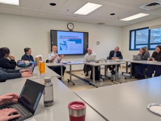 A group of six people in a meeting room sit around a conference table with laptops. A presentation screen shows a slide with a chart. The mood is focused.
