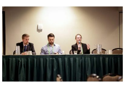 Three people are seated at a panel table. The person on the right is speaking into a microphone. The setting is a conference room with a formal tone.