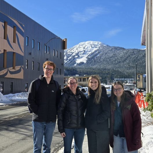 Four people wearing winter jackets stand smiling on a snowy street. A modern building and a snow-capped mountain are visible under a clear blue sky.