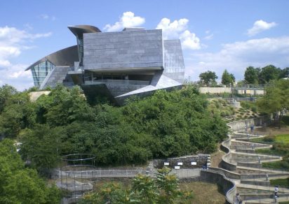 Futuristic building with angular design perched on a lush green hill. A winding path leads through landscaped gardens, under a partly cloudy sky.