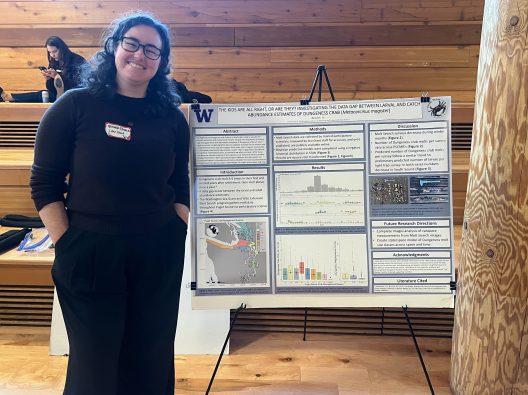 A person stands smiling next to a detailed scientific poster presentation on Dungeness crab abundance estimates. They are in a warmly lit room.