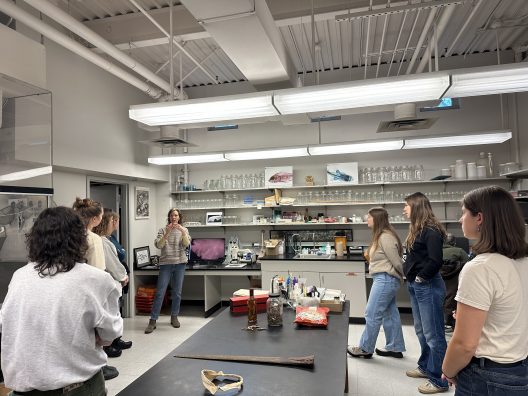 A group of people in a lab observe a person holding up a sample. The room is filled with scientific equipment and glassware, creating an educational atmosphere.