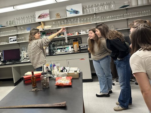 A woman holds up a specimen as a group of students lean in closely, appearing curious. They're in a lab with shelves of jars and scientific equipment.
