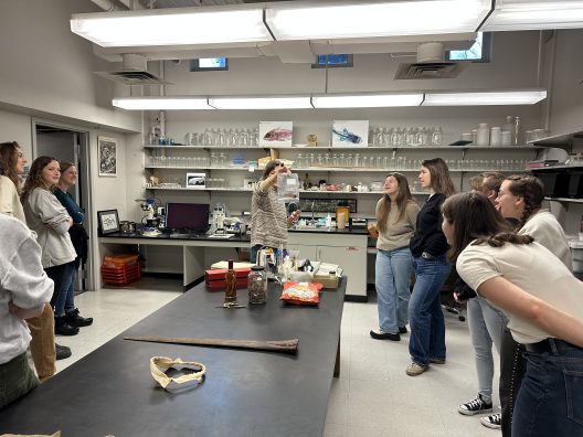 A group of people in a lab watches a woman holding up an object. Shelves with scientific equipment line the walls. The scene is educational and focused.