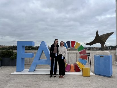 Two people stand smiling in front of a large "FAO" sign with a colorful circular design, set outdoors under a cloudy sky with a sculpture in the background.