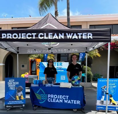 Two people stand under a "Project Clean Water" tent at an outdoor event. The booth is set with educational materials and a sign encouraging clean water practices.
