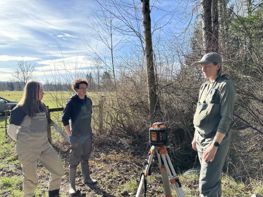 Three people stand outdoors, engaged near a tripod and equipment, surrounded by leafless trees and a grassy field under a bright, partly cloudy sky.