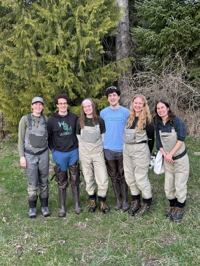 A group of six people stands outdoors in waders and boots, smiling in front of a backdrop of lush evergreen trees, conveying a cheerful tone.