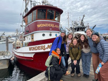 A group of seven smiling young women pose on a dock beside a boat named "Wonderland." They appear relaxed and happy, dressed warmly against a cloudy sky.