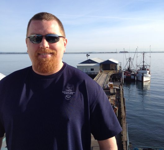 A man with a beard wearing sunglasses and a dark blue shirt stands smiling on a pier. Behind him, a tranquil sea, fishing boats, and a distant ship.