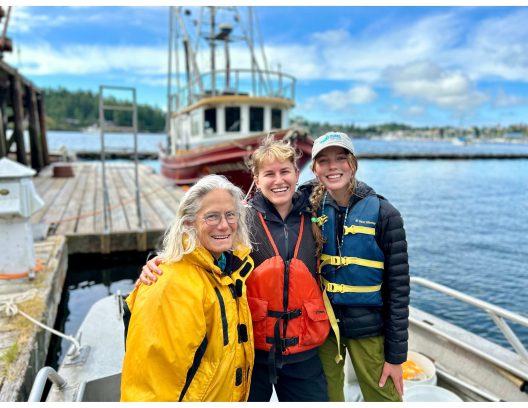 Three smiling people in colorful jackets stand on a boat near a dock, with a wooden ship in the background. The scene conveys joy and camaraderie.