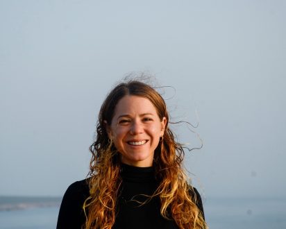 Smiling woman with wavy hair stands in front of a calm, hazy seascape. She wears a black turtleneck, conveying a serene and content mood.