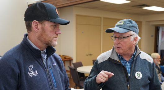 Two men in casual outdoor attire and caps engage in a serious conversation indoors. The setting appears to be a community meeting room.