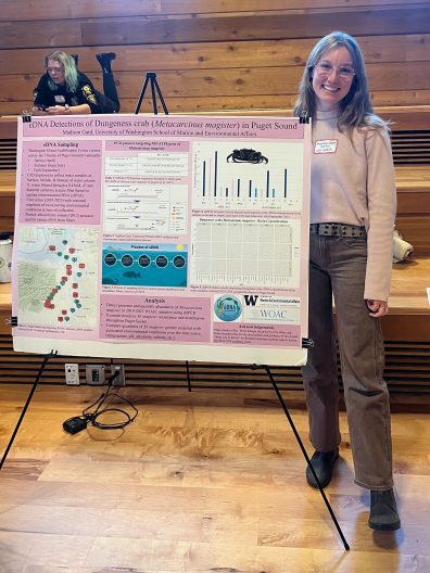 A woman smiles beside a scientific poster on the eDNA detection of Dungeness crabs in Puget Sound. The poster features maps, charts, and text. A person is seated casually on wooden bleachers in the background, creating a relaxed atmosphere.