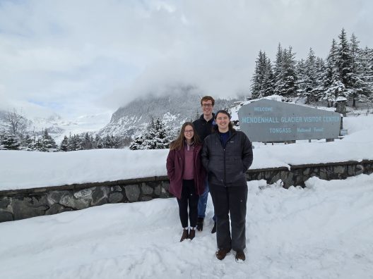 Three people stand smiling in front of the Mendenhall Glacier Visitor Center sign, surrounded by snow-covered trees and mountains under a cloudy sky.