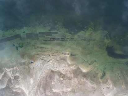 Aerial view of a tranquil shoreline with shallow, clear water revealing submerged structures. A small boat rests on the sandy beach under soft, overcast light.