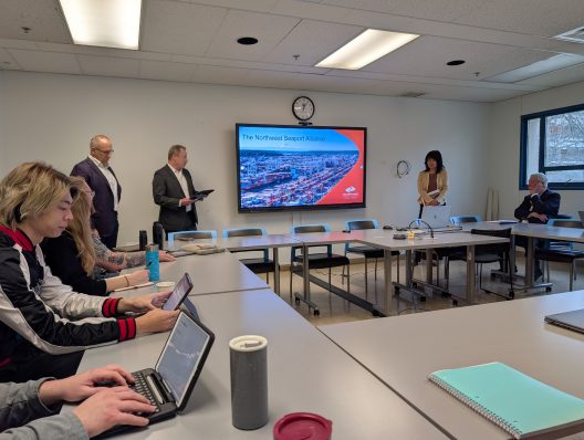 A meeting room with a presentation slide titled "The Northwest Seaport Alliance" displayed on a screen. Four people sit on the left with laptops, while three stand on the right near the screen, engaged in discussion. The atmosphere is professional.