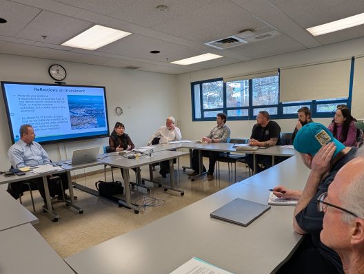 A group of people sitting around a U-shaped table in a meeting room with a large screen showing "Reflections on Investment." The atmosphere is focused and engaged.