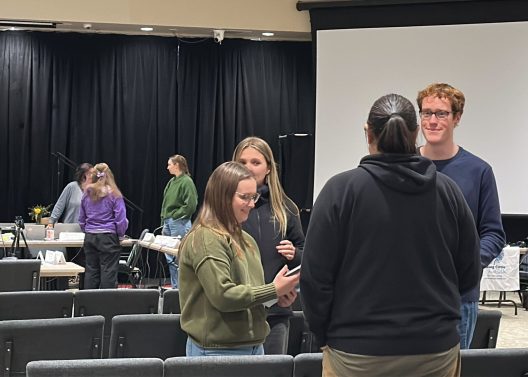 Four people are casually chatting in a room with chairs, a stage, and a white projector screen in the background. The atmosphere is relaxed and friendly.