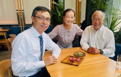 Three people, two men in white shirts and a woman in a floral blouse, smile at a restaurant table with a small wooden tray of desserts in front.
