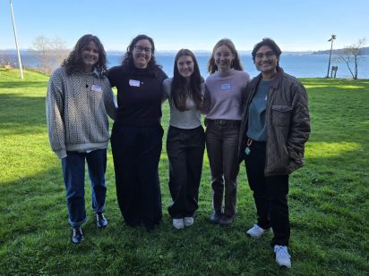 Five people stand closely together, smiling with arms linked, on a sunny grass field by a serene lake. They wear casual, warm clothes and name tags.