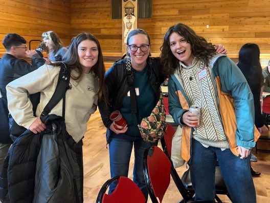 Three people smiling at the camera in a wooden room with others in the background. Each is wearing casual attire and holding jackets, exuding a cheerful vibe.