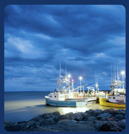 Fishing boats docked at a rocky shore under a dramatic, cloudy twilight sky. The bright lights on the boats contrast with the dark blue hues above.