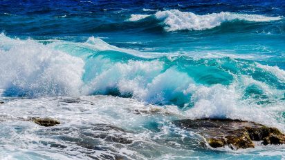 Turquoise waves crash over rocks, under a deep blue sea and sky. The scene conveys a sense of dynamic energy and natural beauty.
