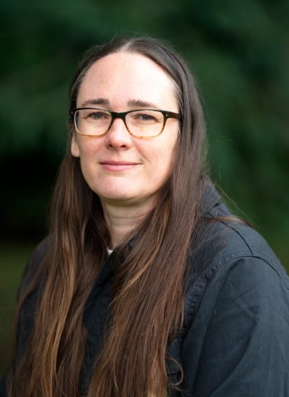 A woman with long brown hair and glasses, wearing a dark jacket, stands outdoors against a blurred green background, smiling softly at the camera.