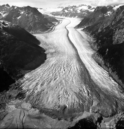 A vast glacier flows between rugged, snow-capped mountains, creating a textured river of ice. The scene conveys majesty and solitude in a remote wilderness.