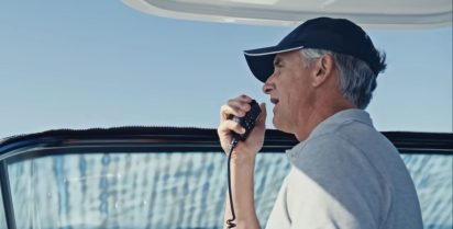 An older man in a cap speaks into a radio microphone on a boat, with a blue sky and water visible in the background, conveying a focused atmosphere.