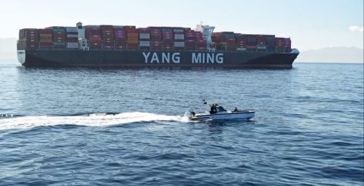 A large container ship labeled "Yang Ming" floats on calm blue waters under clear skies, with a small motorboat speeding by in the foreground.