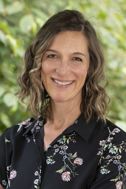 Smiling woman with wavy brown hair wearing a black floral shirt in front of a blurred green foliage background, conveying a warm, cheerful tone.