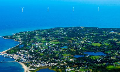 Aerial view of a coastal town surrounded by lush greenery and patches of blue ponds. Wind turbines stand in the distant ocean under a clear blue sky.