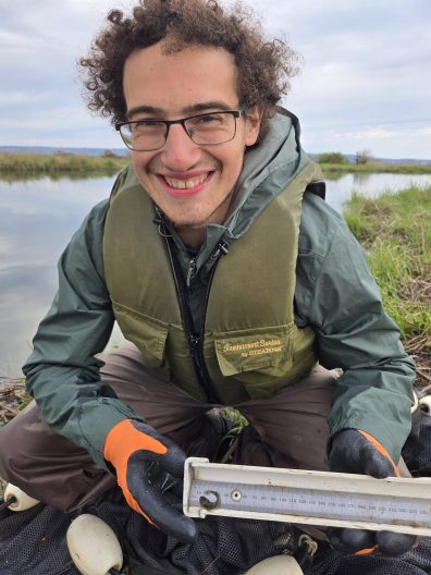 Man in a green jacket and life vest, smiling, holds a ruler over a small fish by a riverside. He wears orange gloves, conveying a cheerful outdoor scene.