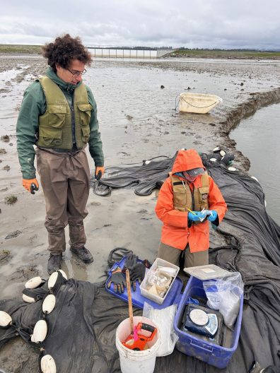 Two people in waterproof gear stand on a muddy shoreline. One, in an orange raincoat, examines a small object; the other observes. Nearby equipment suggests scientific fieldwork.