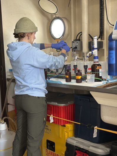 A person in a hoodie and beanie, wearing gloves, works at a lab bench on a ship, surrounded by bottles and equipment, under a porthole's natural light.
