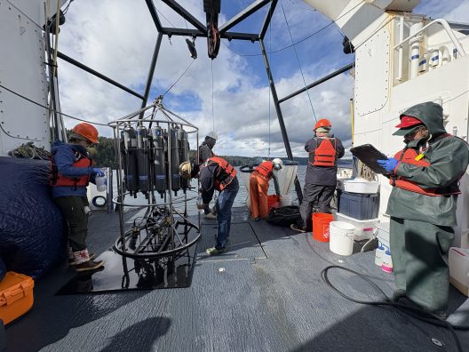 People on a research vessel wearing protective gear work with oceanographic equipment under a cloudy sky, focusing on scientific tasks.